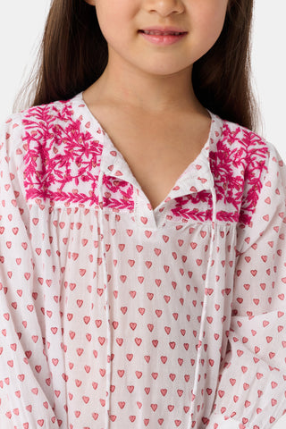 A young girl smiles in the Roller Rabbit Kids Hearts Serafina Tunic, a white cotton dress featuring pink heart patterns, chest embroidery, and tassels on the hem and neckline. The background is plain white.