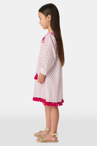 A young girl smiles in the Roller Rabbit Kids Hearts Serafina Tunic, a white cotton dress featuring pink heart patterns, chest embroidery, and tassels on the hem and neckline. The background is plain white.