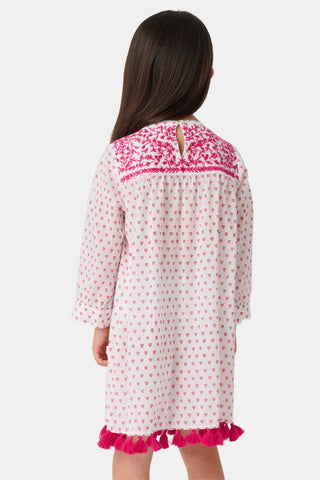 A young girl smiles in the Roller Rabbit Kids Hearts Serafina Tunic, a white cotton dress featuring pink heart patterns, chest embroidery, and tassels on the hem and neckline. The background is plain white.
