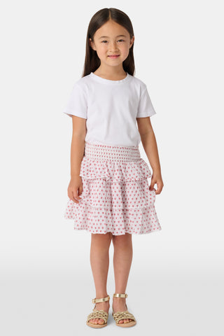A person wears a white short-sleeve top and the Kids Hearts Sami Skirt, a white ruffle mini skirt with pink heart print, standing with hands behind their back against a light background.