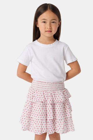 A person wears a white short-sleeve top and the Kids Hearts Sami Skirt, a white ruffle mini skirt with pink heart print, standing with hands behind their back against a light background.