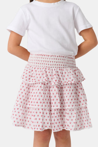A person wears a white short-sleeve top and the Kids Hearts Sami Skirt, a white ruffle mini skirt with pink heart print, standing with hands behind their back against a light background.