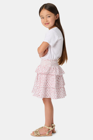 A person wears a white short-sleeve top and the Kids Hearts Sami Skirt, a white ruffle mini skirt with pink heart print, standing with hands behind their back against a light background.