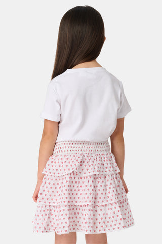 A person wears a white short-sleeve top and the Kids Hearts Sami Skirt, a white ruffle mini skirt with pink heart print, standing with hands behind their back against a light background.