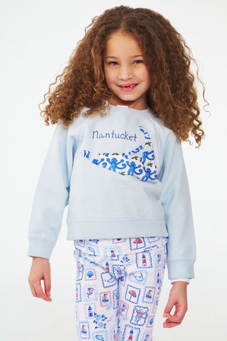 A young girl with curly hair smiles while wearing the Roller Rabbit Kids ACK Applique Crewneck and white pants with a blue and red nautical print, standing against a plain background.