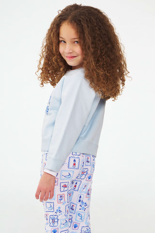 A young girl with curly hair smiles while wearing the Roller Rabbit Kids ACK Applique Crewneck and white pants with a blue and red nautical print, standing against a plain background.