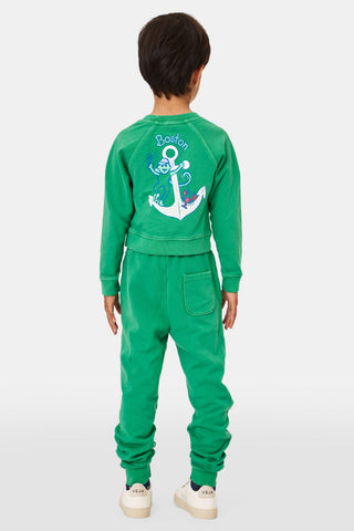 A young child faces forward in Roller Rabbit Kids Boston Monkey Sweatpants, paired with a matching green sweatshirt featuring playful graphics and white sneakers, set against a plain white background.
