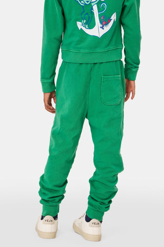 A young child faces forward in Roller Rabbit Kids Boston Monkey Sweatpants, paired with a matching green sweatshirt featuring playful graphics and white sneakers, set against a plain white background.