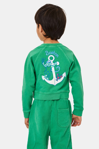 A child faces forward, head partially out of frame, wearing the Roller Rabbit Kids Boston Monkey Crewneck— a green sweatshirt and pants set with a playful cartoon monkey graphic on the upper left.