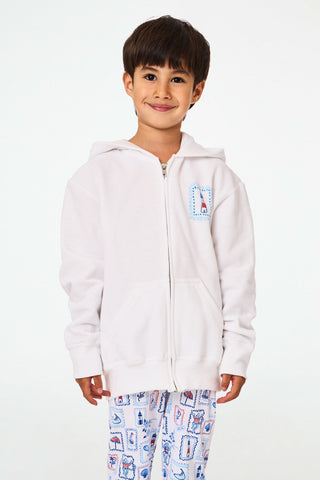 A child is wearing the Roller Rabbit Kids ACK Zip Up hoodie in white, crafted from lightweight French terry and showcasing blue and red nautical designs—including an anchor, shell, and sailboat—on the back. The child is paired with patterned white pants and is turned away to highlight the hoodie’s island-inspired details.