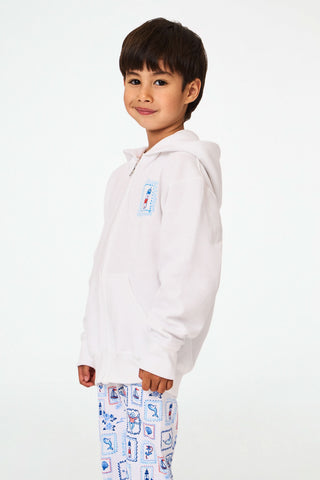 A child is wearing the Roller Rabbit Kids ACK Zip Up hoodie in white, crafted from lightweight French terry and showcasing blue and red nautical designs—including an anchor, shell, and sailboat—on the back. The child is paired with patterned white pants and is turned away to highlight the hoodie’s island-inspired details.