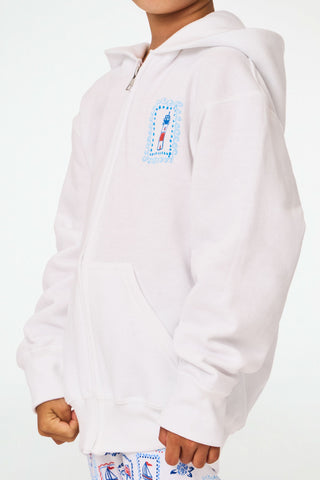 A child is wearing the Roller Rabbit Kids ACK Zip Up hoodie in white, crafted from lightweight French terry and showcasing blue and red nautical designs—including an anchor, shell, and sailboat—on the back. The child is paired with patterned white pants and is turned away to highlight the hoodie’s island-inspired details.