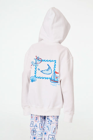 A child is wearing the Roller Rabbit Kids ACK Zip Up hoodie in white, crafted from lightweight French terry and showcasing blue and red nautical designs—including an anchor, shell, and sailboat—on the back. The child is paired with patterned white pants and is turned away to highlight the hoodie’s island-inspired details.