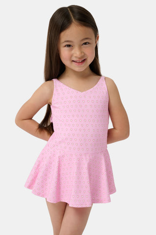 A young girl with long brown hair smiles as she wears the Roller Rabbit Kids Hearts Dory One Piece, a sleeveless pink dress with a flared, ruffled skirt and heart print, standing with her hands behind her back against a light background.