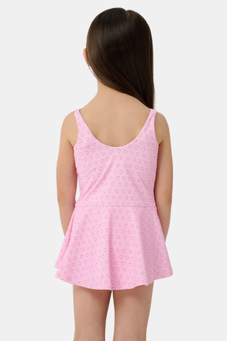 A young girl with long brown hair smiles as she wears the Roller Rabbit Kids Hearts Dory One Piece, a sleeveless pink dress with a flared, ruffled skirt and heart print, standing with her hands behind her back against a light background.