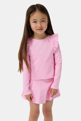 A young girl with light brown hair stands on a sandy beach, smiling in the Roller Rabbit Kids Hearts Ruffle Rashguard. The long-sleeve rashguard features a heart print. Water, trees, and a dock are visible in the background.
