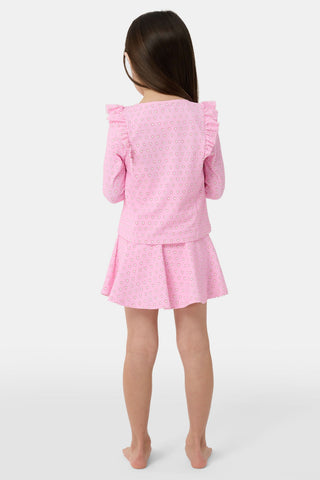 A young girl with light brown hair stands on a sandy beach, smiling in the Roller Rabbit Kids Hearts Ruffle Rashguard. The long-sleeve rashguard features a heart print. Water, trees, and a dock are visible in the background.