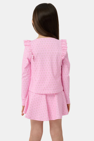 A young girl with light brown hair stands on a sandy beach, smiling in the Roller Rabbit Kids Hearts Ruffle Rashguard. The long-sleeve rashguard features a heart print. Water, trees, and a dock are visible in the background.