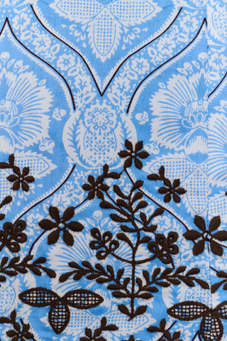 A woman wearing the Roller Rabbit Lakai Asher Dress—a light blue patterned cotton batiste dress with dark lace trim—walks on grass near a cream-colored house with green shutters, partially visible.