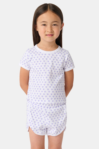 A young child with long brown hair smiles, wearing the Roller Rabbit Kids Hearts Playa Play Set—featuring a small purple heart pattern—hands behind their back, against a plain light background.