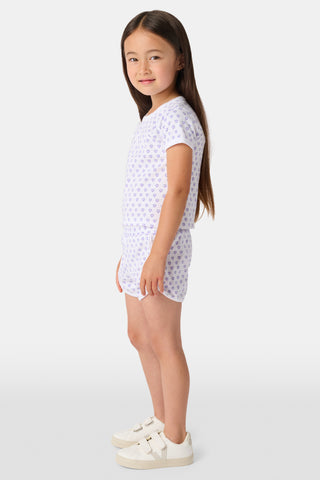 A young child with long brown hair smiles, wearing the Roller Rabbit Kids Hearts Playa Play Set—featuring a small purple heart pattern—hands behind their back, against a plain light background.