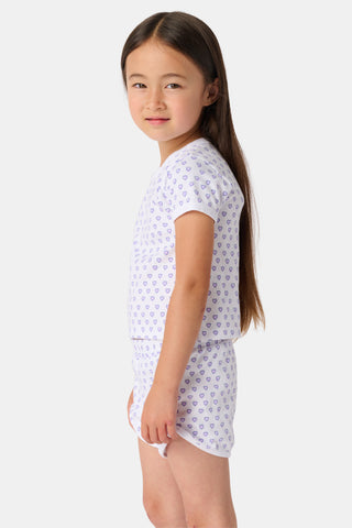 A young child with long brown hair smiles, wearing the Roller Rabbit Kids Hearts Playa Play Set—featuring a small purple heart pattern—hands behind their back, against a plain light background.