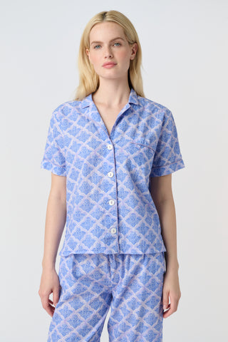 A person stands barefoot against a plain background, wearing the Roller Rabbit Jemina Liza Set—a light blue cotton voile loungewear set with a geometric pattern, featuring a short-sleeve button-up shirt and loose-fitting pants.