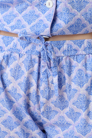 A person stands barefoot against a plain background, wearing the Roller Rabbit Jemina Liza Set—a light blue cotton voile loungewear set with a geometric pattern, featuring a short-sleeve button-up shirt and loose-fitting pants.