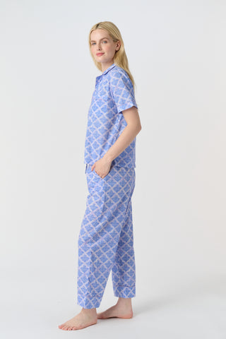 A person stands barefoot against a plain background, wearing the Roller Rabbit Jemina Liza Set—a light blue cotton voile loungewear set with a geometric pattern, featuring a short-sleeve button-up shirt and loose-fitting pants.