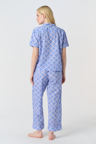 A person stands barefoot against a plain background, wearing the Roller Rabbit Jemina Liza Set—a light blue cotton voile loungewear set with a geometric pattern, featuring a short-sleeve button-up shirt and loose-fitting pants.