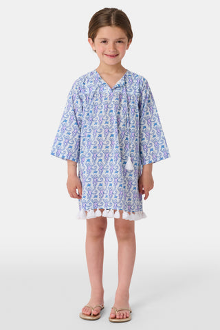 A young girl with brown hair tied back smiles in a Roller Rabbit Kids Monkey Serafina Tunic—a white and blue patterned cotton voile dress with long sleeves, white tassels at the hem, and flip-flops—against a plain white background.