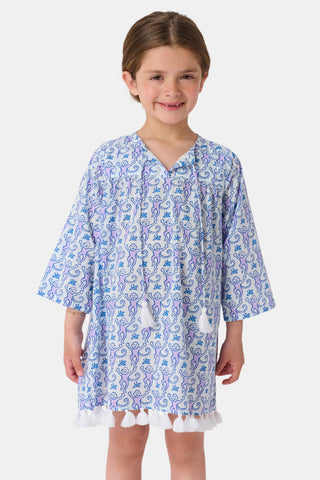 A young girl with brown hair tied back smiles in a Roller Rabbit Kids Monkey Serafina Tunic—a white and blue patterned cotton voile dress with long sleeves, white tassels at the hem, and flip-flops—against a plain white background.