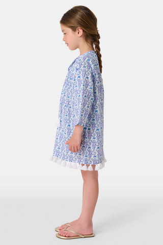A young girl with brown hair tied back smiles in a Roller Rabbit Kids Monkey Serafina Tunic—a white and blue patterned cotton voile dress with long sleeves, white tassels at the hem, and flip-flops—against a plain white background.