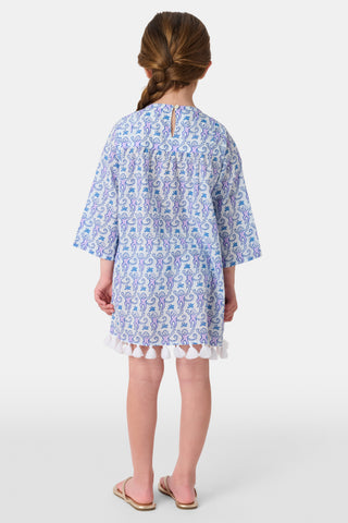 A young girl with brown hair tied back smiles in a Roller Rabbit Kids Monkey Serafina Tunic—a white and blue patterned cotton voile dress with long sleeves, white tassels at the hem, and flip-flops—against a plain white background.