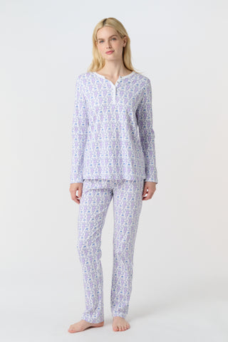 A woman stands barefoot against a white background, wearing Roller Rabbit's Monkey Pajamas—long-sleeved, buttoned 100 Pima Cotton pajamas featuring a matching top and bottom with a light purple and white floral print.