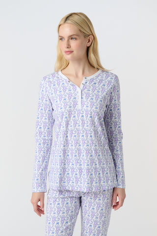 A woman stands barefoot against a white background, wearing Roller Rabbit's Monkey Pajamas—long-sleeved, buttoned 100 Pima Cotton pajamas featuring a matching top and bottom with a light purple and white floral print.