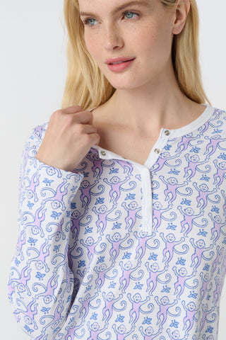 A woman stands barefoot against a white background, wearing Roller Rabbit's Monkey Pajamas—long-sleeved, buttoned 100 Pima Cotton pajamas featuring a matching top and bottom with a light purple and white floral print.