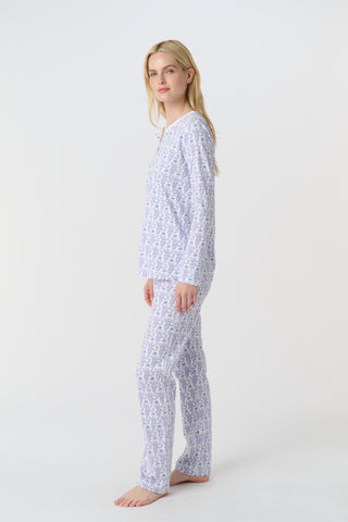 A woman stands barefoot against a white background, wearing Roller Rabbit's Monkey Pajamas—long-sleeved, buttoned 100 Pima Cotton pajamas featuring a matching top and bottom with a light purple and white floral print.