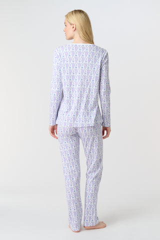 A woman stands barefoot against a white background, wearing Roller Rabbit's Monkey Pajamas—long-sleeved, buttoned 100 Pima Cotton pajamas featuring a matching top and bottom with a light purple and white floral print.