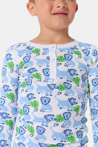 A young child with curly hair smiles barefoot against a plain white background while wearing Kids Leo The Lion Pajamas, crafted from soft Pima cotton.