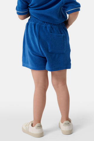A child wears the Roller Rabbit Kids Monkey Embroidery Hanni Short, navy terry shorts with a monkey embroidered on the left leg, paired with a matching blue top and white velcro sneakers against a plain white background.