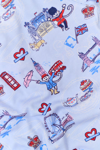 A young child with curly hair stands barefoot against a plain white background, wearing Roller Rabbit’s Kids London Monkey Pajamas—light blue long-sleeve pajamas adorned with playful, colorful patterns featuring guards, clocks, and iconic British landmarks.