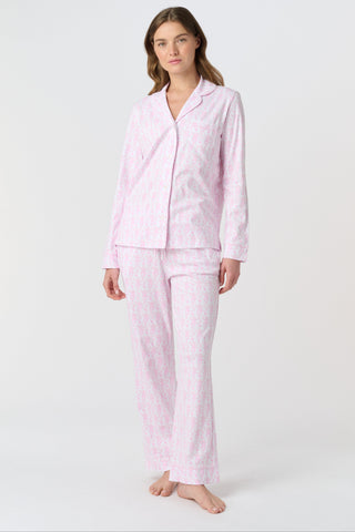 A woman with long, wavy brown hair stands barefoot against a white background, wearing the Lovely Monkey Long Sleeve Polo Pajamas in light pink with a subtle floral pattern—a comfy button-up top and matching pants perfect for relaxing nights.