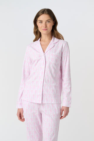 A woman with long, wavy brown hair stands barefoot against a white background, wearing the Lovely Monkey Long Sleeve Polo Pajamas in light pink with a subtle floral pattern—a comfy button-up top and matching pants perfect for relaxing nights.