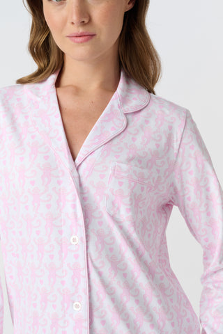 A woman with long, wavy brown hair stands barefoot against a white background, wearing the Lovely Monkey Long Sleeve Polo Pajamas in light pink with a subtle floral pattern—a comfy button-up top and matching pants perfect for relaxing nights.