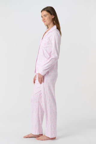 A woman with long, wavy brown hair stands barefoot against a white background, wearing the Lovely Monkey Long Sleeve Polo Pajamas in light pink with a subtle floral pattern—a comfy button-up top and matching pants perfect for relaxing nights.