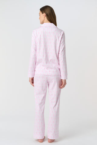 A woman with long, wavy brown hair stands barefoot against a white background, wearing the Lovely Monkey Long Sleeve Polo Pajamas in light pink with a subtle floral pattern—a comfy button-up top and matching pants perfect for relaxing nights.