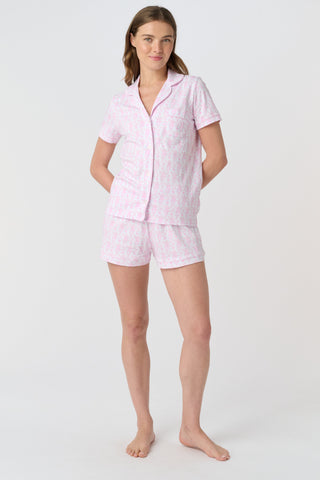 A woman with long, wavy brown hair stands barefoot against a white background, wearing the Lovely Monkey Long Sleeve Polo Pajamas in light pink with a subtle floral pattern—a comfy button-up top and matching pants perfect for relaxing nights.