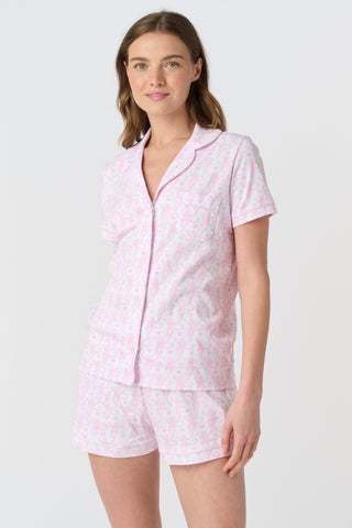 A woman with long, wavy brown hair stands barefoot against a white background, wearing the Lovely Monkey Long Sleeve Polo Pajamas in light pink with a subtle floral pattern—a comfy button-up top and matching pants perfect for relaxing nights.