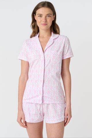 A woman with long, wavy brown hair stands barefoot against a white background, wearing the Lovely Monkey Long Sleeve Polo Pajamas in light pink with a subtle floral pattern—a comfy button-up top and matching pants perfect for relaxing nights.
